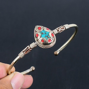 May include: A silver bangle bracelet featuring a teardrop pendant with turquoise and red stones. The pendant is intricately detailed and has a rope-like border.
