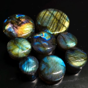 May include: A collection of polished labradorite stones in various sizes. The stones display iridescent flashes of blue, green, and gold against a dark background. These natural gemstones are used for jewelry and decorative purposes.