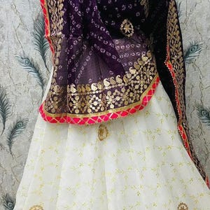 Könnte beinhalten: Traditionelles indisches Outfit mit einem weißen Rock mit goldener Stickerei und einem passenden lila-goldenen Schal. Der Schal hat einen rosa Rand. Das Outfit wird auf einer Schaufensterpuppe präsentiert.