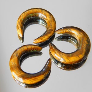 Peut inclure: Trois écarteurs d'oreille en forme de croissant avec un motif œil de tigre. Les écarteurs sont de couleur marron chaud avec des accents marron foncé et verts. La surface lisse et polie reflète la lumière.