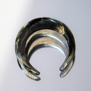 Peut inclure: Une bague en verre transparent avec une surface lisse et arrondie. La bague est en forme de croissant de lune et a une finition polie.