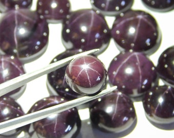 May include: A close-up of a group of dark purple star-shaped gemstones. The gemstones are round and have a shiny, polished finish. One gemstone is being held by tweezers.