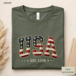 May include: An army green t-shirt featuring the embroidered word "USA" in red, white, and blue, with a stars and stripes design. The text "EST. 1776" is embroidered below. The t-shirt is folded.