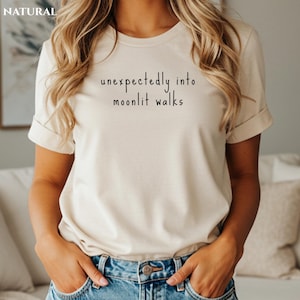 May include: A beige t-shirt with the text "unexpectedly into moonlit walks" in black script. The shirt has short sleeves and a crew neck. The word "NATURAL" is in the upper left corner.
