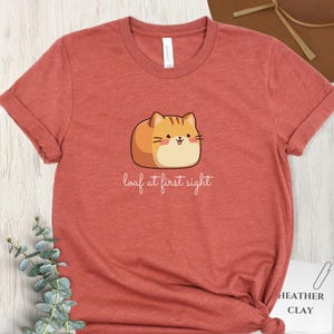 May include: A heather clay-colored t-shirt featuring a cartoon cat illustration. The cat is orange and tan, with a smiling face. The text "loaf at first sight" is below the cat. The shirt has rolled-up sleeves.