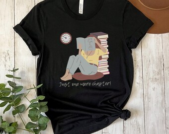 May include: Black t-shirt featuring a reading illustration with a clock and books. The text "Just one more chapter!" is printed below the image. The design shows a person sitting on a cushion, engrossed in a book.