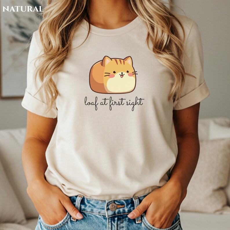 May include: A beige t-shirt featuring a cartoon cat designed to look like a loaf of bread. The cat is orange and tan with a smiling face. The text "loaf at first sight" is written below the cat in a handwritten font.