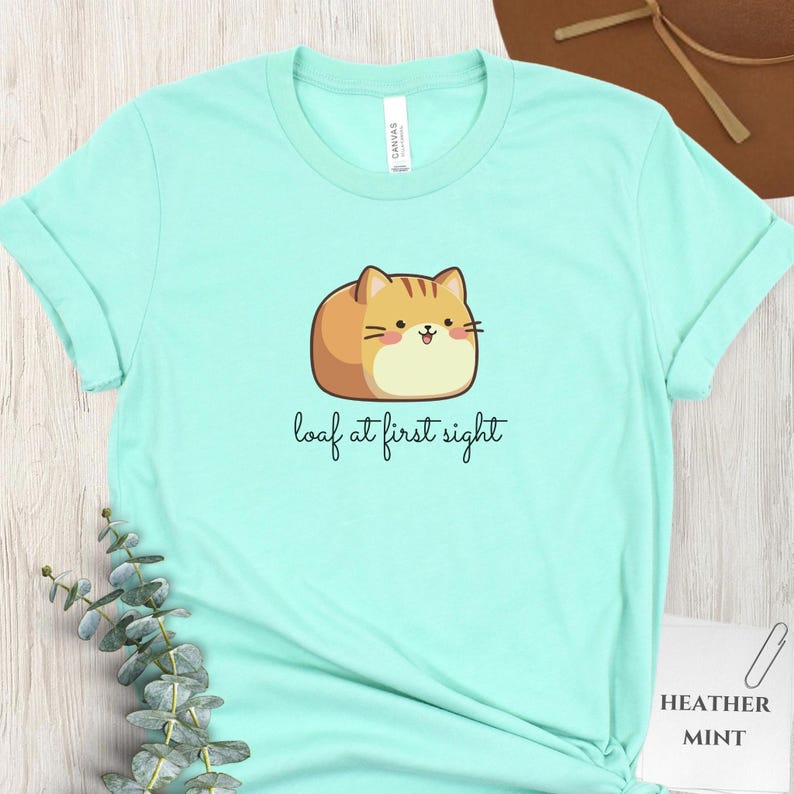 May include: Mint green t-shirt with a cartoon cat illustration. The cat is brown and tan, shaped like a loaf of bread, with the text "loaf at first sight" in a handwritten font. The shirt is made of a soft material.