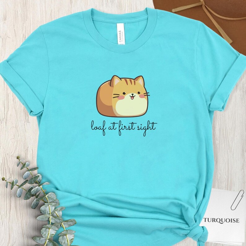 May include: Turquoise t-shirt with a cartoon cat illustration and the phrase "loaf at first sight". The cat is tan and cream-colored with a smiling face. The shirt is made of a soft material.