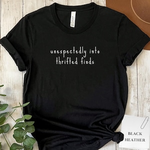 May include: Black t-shirt with the white text "unexpectedly into thrifted finds." The shirt is made of a soft material and has short sleeves. The tag on the bottom right corner says "Black Heather."