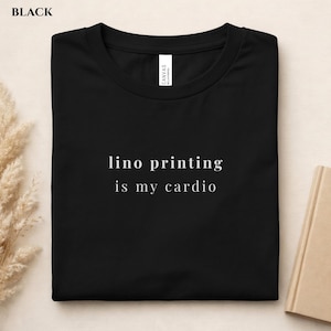 May include: Black t-shirt with the text "lino printing is my cardio" in white. The shirt is folded and has a tag that says "Canvas Bella+Canvas". The shirt is on a light brown surface.