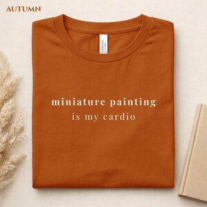 May include: An orange t-shirt with the text "miniature painting is my cardio" in white. The shirt is folded and has a tag that says "CANVAS". The word "AUTUMN" is in the top left corner.