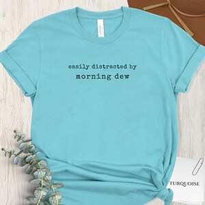 Pode incluir: Camiseta turquesa com a frase "easily distracted by morning dew" em preto. A camiseta tem gola redonda e é feita de um tecido macio. A palavra "TURQUOISE" está impressa em um cartão branco.
