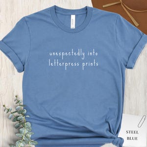 May include: Steel blue t-shirt with rolled-up sleeves. The shirt features the text "unexpectedly into letterpress prints" in white. A tag on the shirt reads "CANVAS". A small card in the bottom right corner reads "STEEL BLUE".