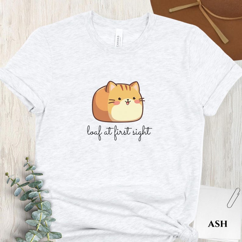 May include: Light gray t-shirt featuring a cartoon cat designed to look like a loaf of bread. The cat is orange and tan with a smiling face. The text "loaf at first sight" is written below the cat in a cursive font.