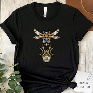 May include: Black t-shirt featuring a detailed, steampunk-style dragonfly and spider design in gold, blue, and white. The insect illustrations are intricate and ornate. The words "BLACK HEATHER" are printed on a white card.