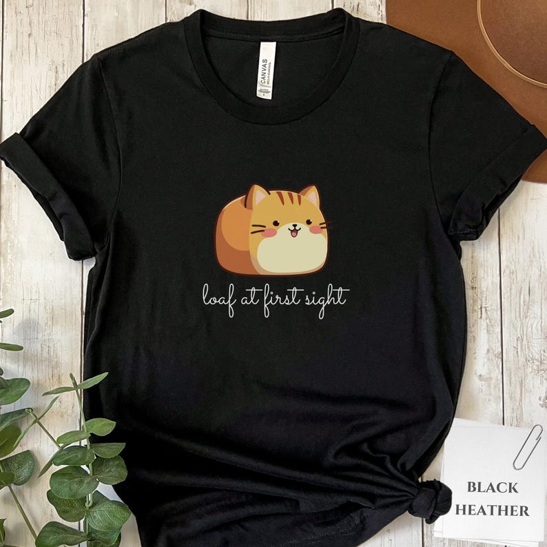 May include: Black t-shirt featuring a cartoon cat designed to look like a loaf of bread. The cat is brown and tan with a smiling face. The text "loaf at first sight" is written below the cat in a cursive font.