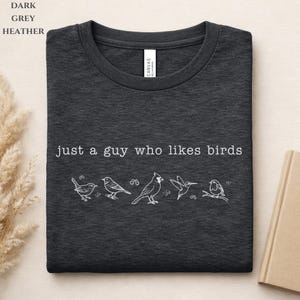 May include: Dark grey heather t-shirt featuring the phrase "just a guy who likes birds" and a white illustration of five birds. The shirt is folded.