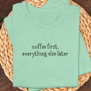 May include: A light green t-shirt with the text "coffee first, everything else later" in black. The shirt is made of a soft material and has a crew neck. The shirt is folded on a woven, light brown surface.