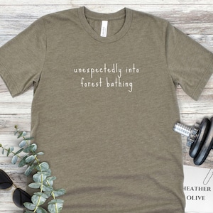 May include: Olive green t-shirt with the text "unexpectedly into forest bathing" in white script. The shirt is made of a soft material and has a crew neck. The shirt is on a light wood surface.