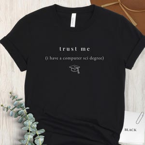 Può includere: T-shirt nero con testo bianco "trust me" sopra "(i have a computer sci degree)". Sotto c'è una piccola grafica di un cappello di laurea. La maglietta è a maniche corte e girocollo.
