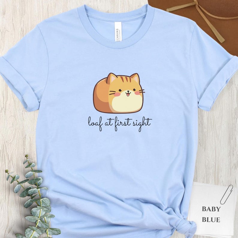 May include: Baby blue t-shirt featuring a cartoon cat designed to look like a loaf of bread. The cat is tan and orange with a smiling face. The text "loaf at first sight" is written below the cat.