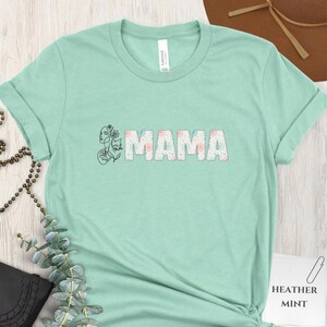 Può includere: T-shirt verde menta con la scritta "MAMA" in un motivo floreale. Un disegno a tratto di una donna e un gatto è sul lato sinistro della parola. La maglietta è realizzata in un materiale morbido.