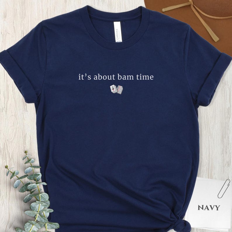 May include: Navy blue t-shirt with rolled-up sleeves. The shirt features the text "it's about bam time" in white, along with a small graphic of two dice. The word "NAVY" is printed in white on a small white rectangle.