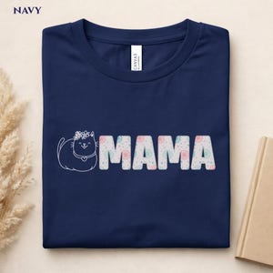 Könnte beinhalten: Marineblaues T-Shirt mit dem Wort "MAMA" in Weiß, verziert mit einem Blumenmuster. Auf der linken Seite befindet sich eine weiße Umrisszeichnung einer Katze mit Blumenkrone. Das T-Shirt ist aus weichem Material.