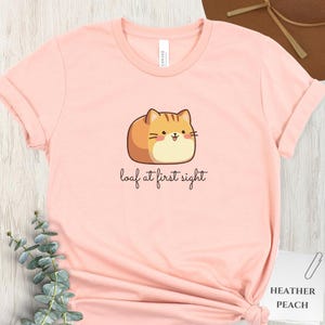May include: A light pink t-shirt with a cartoon cat illustration. The cat is brown and orange, resembling a loaf of bread. The text "loaf at first sight" is written below the cat in a cursive font. The shirt is labeled "Heather Peach".