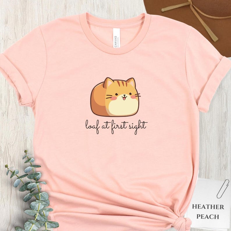 May include: A light pink t-shirt with a cartoon cat illustration. The cat is brown and orange, resembling a loaf of bread. The text "loaf at first sight" is written below the cat in a cursive font. The shirt is labeled "Heather Peach".