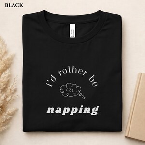 May include: Black t-shirt with the text "I'd rather be napping" in white. The design includes a cloud with "Zzz" inside. The shirt is folded and has a tag that says "Canvas".