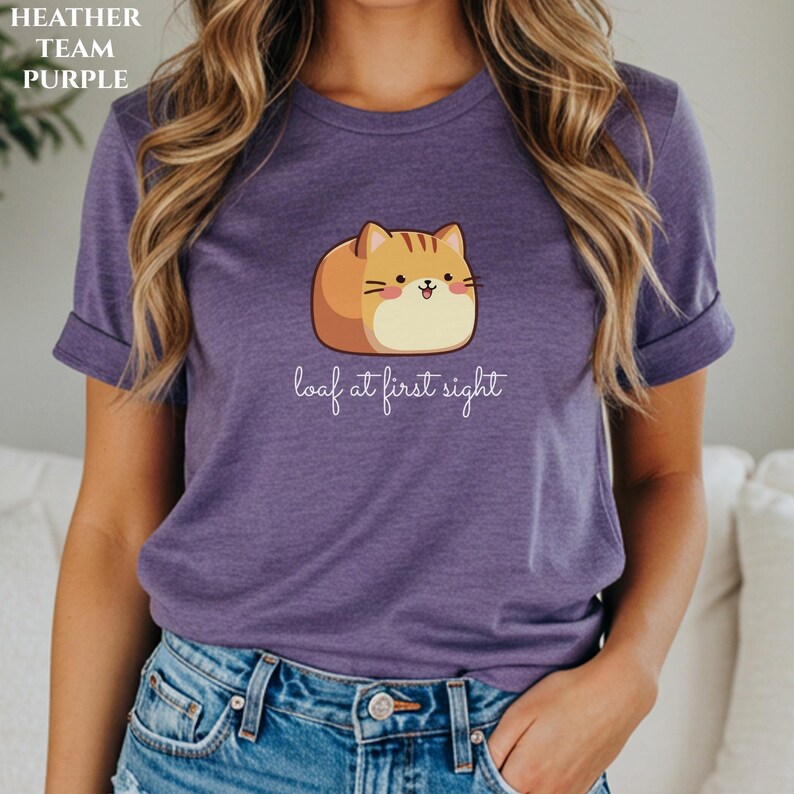 May include: Purple t-shirt with a cartoon cat-loaf graphic. The cat is tan and brown with a smiling face. The text "loaf at first sight" is below the cat. The shirt is heather purple.