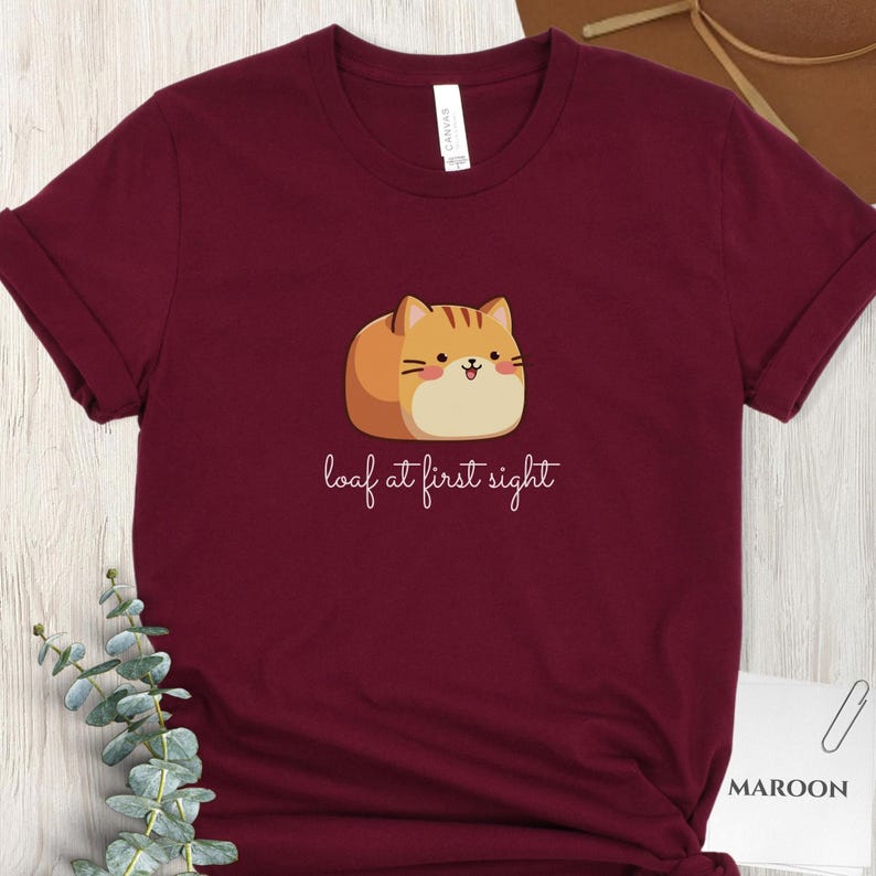 May include: Maroon t-shirt featuring a cartoon cat designed to look like a loaf of bread. The cat is orange and tan with a smiling face. The text "loaf at first sight" is written below the cat in a cursive font.