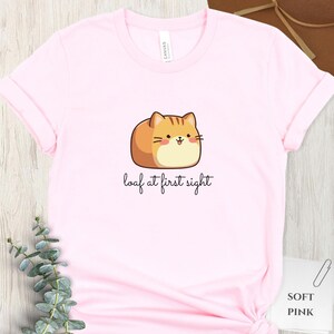 May include: A light pink t-shirt featuring a cartoon cat illustration with the text "loaf at first sight". The cat is brown and tan with a smiling face. The shirt is made of soft material.