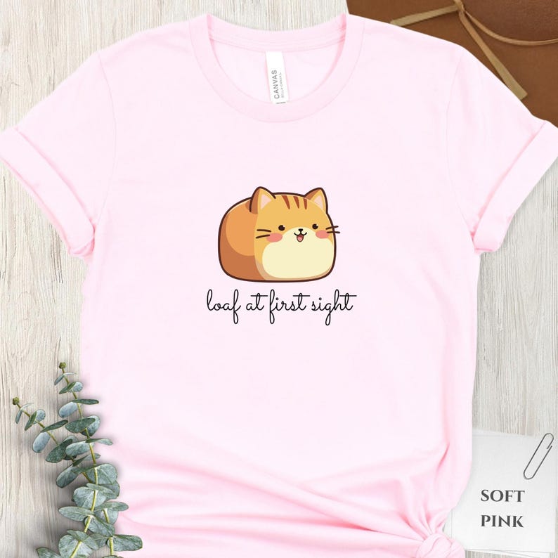 May include: A light pink t-shirt featuring a cartoon cat illustration with the text "loaf at first sight". The cat is brown and tan with a smiling face. The shirt is made of soft material.