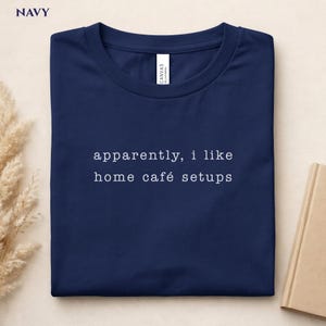 May include: Navy blue t-shirt with the text "apparently, i like home café setups" in white. The shirt is folded and lying flat. The word "NAVY" is in the top left corner.