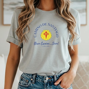 May include: A light gray t-shirt featuring the words "CAMINO DE SANTIAGO" in blue, arched above a yellow scallop shell with a red cross. Below the shell, the text "Buen Camino...Soon!" is written in blue.