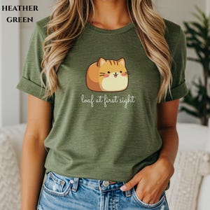 May include: Heather green t-shirt featuring a cartoon cat designed to look like a loaf of bread. The cat is brown and tan with a smiling face. The text "loaf at first sight" is written below the cat in a cursive font.