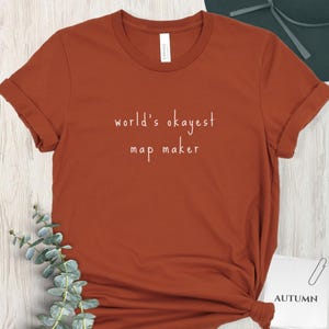 May include: A rust-colored t-shirt with rolled-up sleeves. The text "world's okayest map maker" is printed in white script across the front. The shirt is displayed on a light-colored surface with a sprig of greenery and a card that says "AUTUMN".