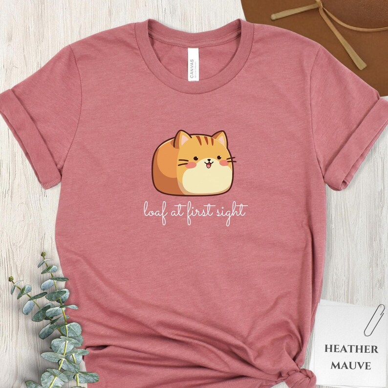 May include: Heather mauve t-shirt with a cartoon cat illustration. The cat is orange and tan, with a smiling face. The text "loaf at first sight" is written below the cat in a cursive font. The shirt is made of a soft material.