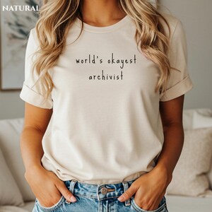 May include: A beige t-shirt with the text "world's okayest archivist" in black. The short-sleeved shirt is made of a soft material and is being worn. The word "NATURAL" is in the top left corner.