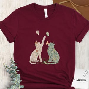 以下が含まれることがあります： 2匹の猫と蝶の気まぐれなデザインが特徴のマルーンTシャツ。1匹の猫が蝶に向かって手を伸ばしています。猫は花柄で装飾されています。「MAROON」の文字が白いラベルに印刷されています。