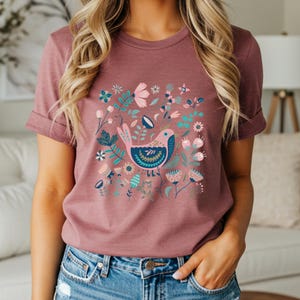 May include: A mauve-colored t-shirt featuring a colorful bird and floral design. The bird is pink and blue, surrounded by flowers in shades of pink, teal, and white. The short-sleeved shirt is made of soft material.