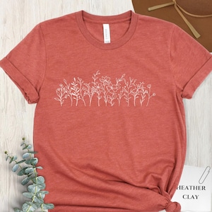 May include: A coral-colored t-shirt with a white floral design across the chest. The shirt has rolled-up sleeves and a crew neck. The tag reads "Canvas". The bottom right corner of the shirt has a tag that reads "Heather Clay".