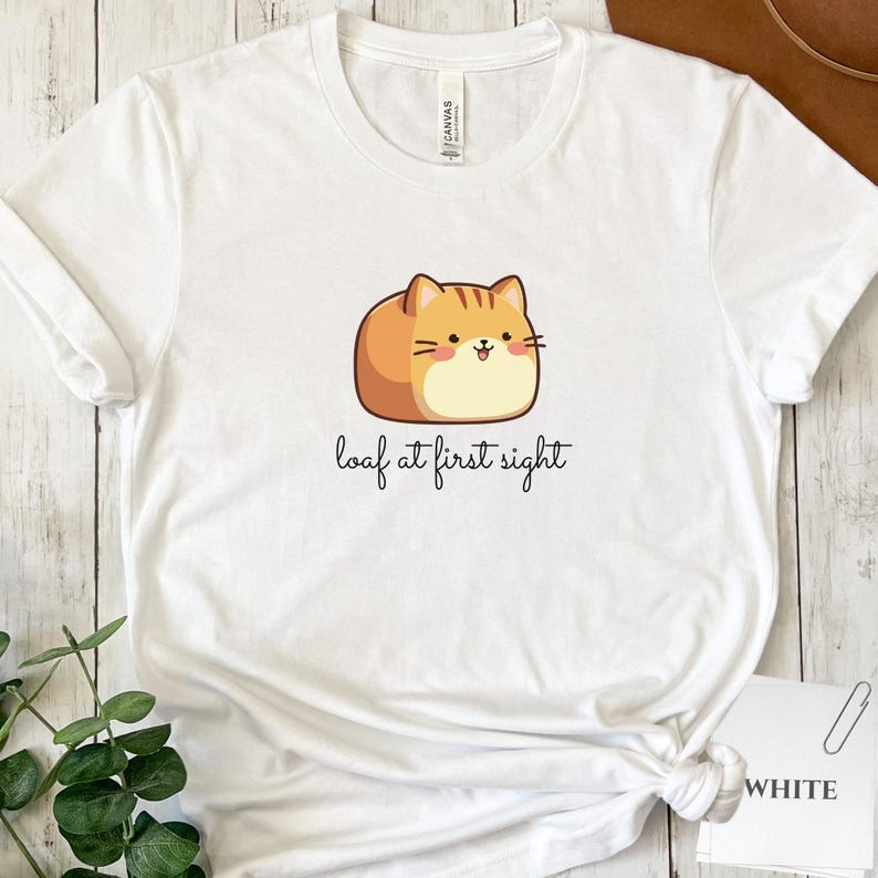 May include: White t-shirt featuring a cartoon cat in a loaf shape, with orange and tan fur and a smiling face. The text "loaf at first sight" is printed below the cat. The shirt is made of a soft material.
