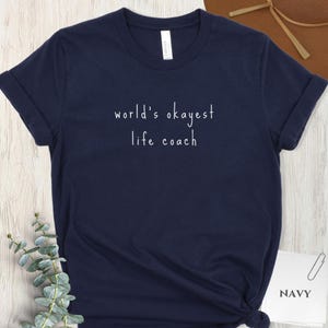 May include: Navy blue t-shirt with the text "world's okayest life coach" in white. The shirt is made of a soft material and has rolled-up sleeves. A small tag on the shirt reads "Canvas".