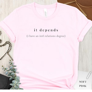 May include: A pale pink t-shirt featuring the words "it depends" and the phrase "(i have an intl relations degree)" in black. The shirt has rolled sleeves and is made of a soft fabric.