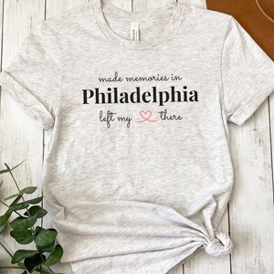Könnte beinhalten: Hellgraues T-Shirt mit dem Text "made memories in Philadelphia left my heart there" in Schwarz und Rosa. Das Shirt ist aus einem weichen Material und hat kurze Ärmel.