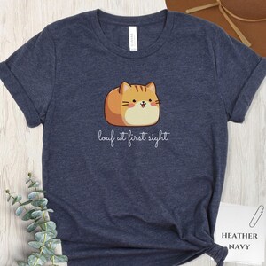 May include: Heather navy t-shirt featuring a cartoon cat designed to look like a loaf of bread. The cat is orange and tan with a smiling face. The text "loaf at first sight" is written below the cat.